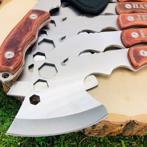 May include: A collection of stainless steel hatchets and multi-tools with wooden handles. The hatchets have a silver blade and a brown wooden handle. Some handles are engraved with names. The tools are arranged on a wooden surface.