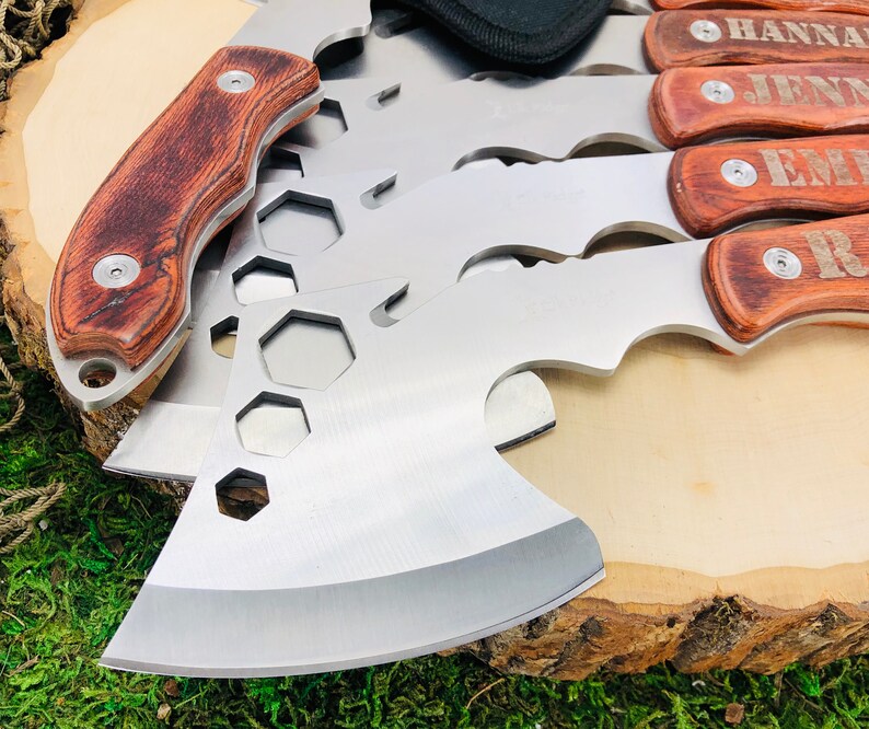 May include: A collection of stainless steel hatchets and multi-tools with wooden handles. The hatchets have a silver blade and a brown wooden handle. Some handles are engraved with names. The tools are arranged on a wooden surface.