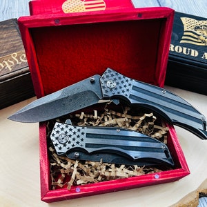 May include: Two black folding knives with American flag patterns on the handles are displayed in a red wooden box with a red velvet lining. The box is sitting on a wooden surface.