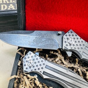 May include: Two silver folding knives with a stars and stripes pattern on the handle. The blade of one knife has an engraved image of a snake with the words "Don't Tread On Me".