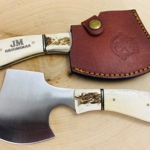 May include: A small axe with a white bone handle and a leather sheath. The axe has a stainless steel blade and a brass accent on the handle. The sheath has a brown leather strap with a brass button. The axe is engraved with the initials "JM" and the word "Groomsman".