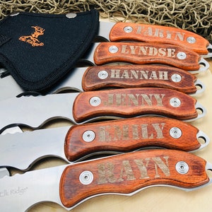 May include: A collection of personalized hunting knives with wooden handles, each engraved with a different name. The blades are silver, and the handles are a rich brown. A black sheath with the "Elk Ridge" logo is visible.