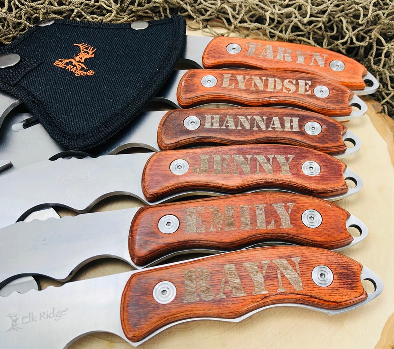 May include: A collection of personalized hunting knives with wooden handles, each engraved with a different name. The blades are silver, and the handles are a rich brown. A black sheath with the "Elk Ridge" logo is visible.