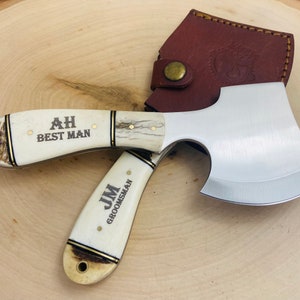 May include: A small hatchet with a polished steel blade and a cream-colored handle. The handle has gold accents and the text "AH BEST MAN" and "JM GROOMSMAN". A brown leather sheath protects the blade.
