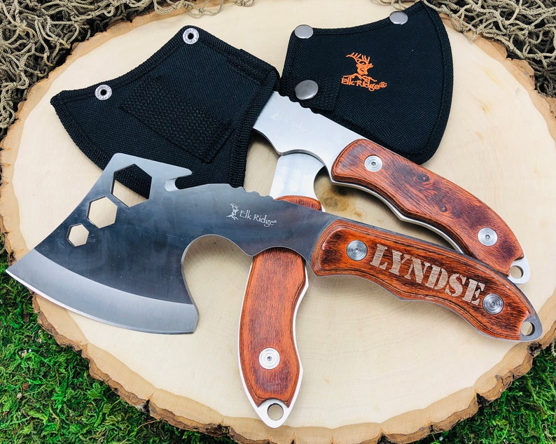 May include: Two axes with wooden handles and stainless steel blades are displayed. One axe has the name "LYNDSE" engraved on the handle. The axes are accompanied by black sheaths. The "Elk Ridge" logo is visible on the blades and sheaths.