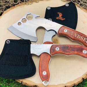 May include: Two stainless steel hatchets and a folding knife with wooden handles. The hatchets have a silver blade and a black sheath. The folding knife has a wooden handle and a silver blade. The text "Elk Ridge" is visible on the blades.