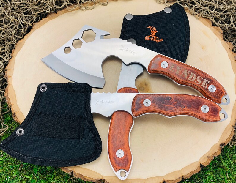 May include: Two stainless steel hatchets and a folding knife with wooden handles. The hatchets have a silver blade and a black sheath. The folding knife has a wooden handle and a silver blade. The text "Elk Ridge" is visible on the blades.
