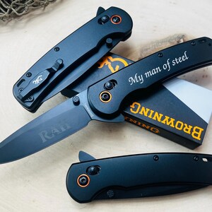 May include: Three black folding knives with orange accents and the text "My man of steel" are displayed on a wooden surface. The knives are from the Browning brand and include a box with the brand name.