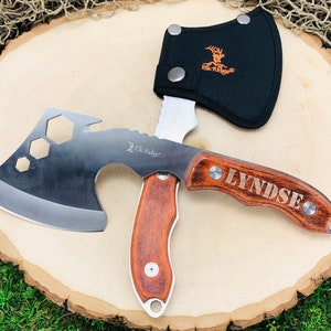 May include: A multi-tool axe with a wooden handle and the name "LYNDSE" engraved on it. The axe head is silver with a cutting edge and hexagonal cutouts. A black sheath with the "Elk Ridge" logo protects the blade.