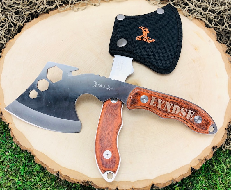 May include: A multi-tool axe with a wooden handle and the name "LYNDSE" engraved on it. The axe head is silver with a cutting edge and hexagonal cutouts. A black sheath with the "Elk Ridge" logo protects the blade.
