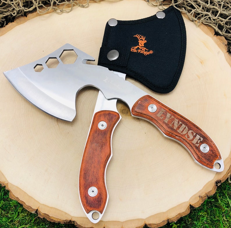 May include: A silver and wood hatchet with a black sheath. The hatchet has a stainless steel blade with a hexagonal cut-out design. The handle is made of wood and has the name "LYNDSE" engraved on it. The black sheath has the "Elk Ridge" logo.