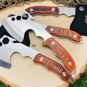 May include: A collection of hatchets with stainless steel blades and wooden handles. The hatchets have a hexagonal design on the blade and are engraved with names. One hatchet is in a black sheath with an orange logo.