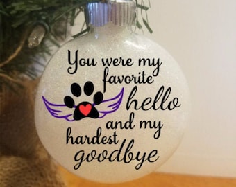Op de afbeelding: Witte glitter kerstbal met een pootprint en vleugels, en de tekst "Je was mijn favoriete hallo en mijn moeilijkste vaarwel".
