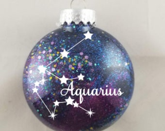 Op de afbeelding: Een paarse en blauwe glazen kerstbal met glitter en een witte omtrek van het sterrenbeeld Waterman. Het woord "Aquarius" is geschreven in witte cursieve letters.