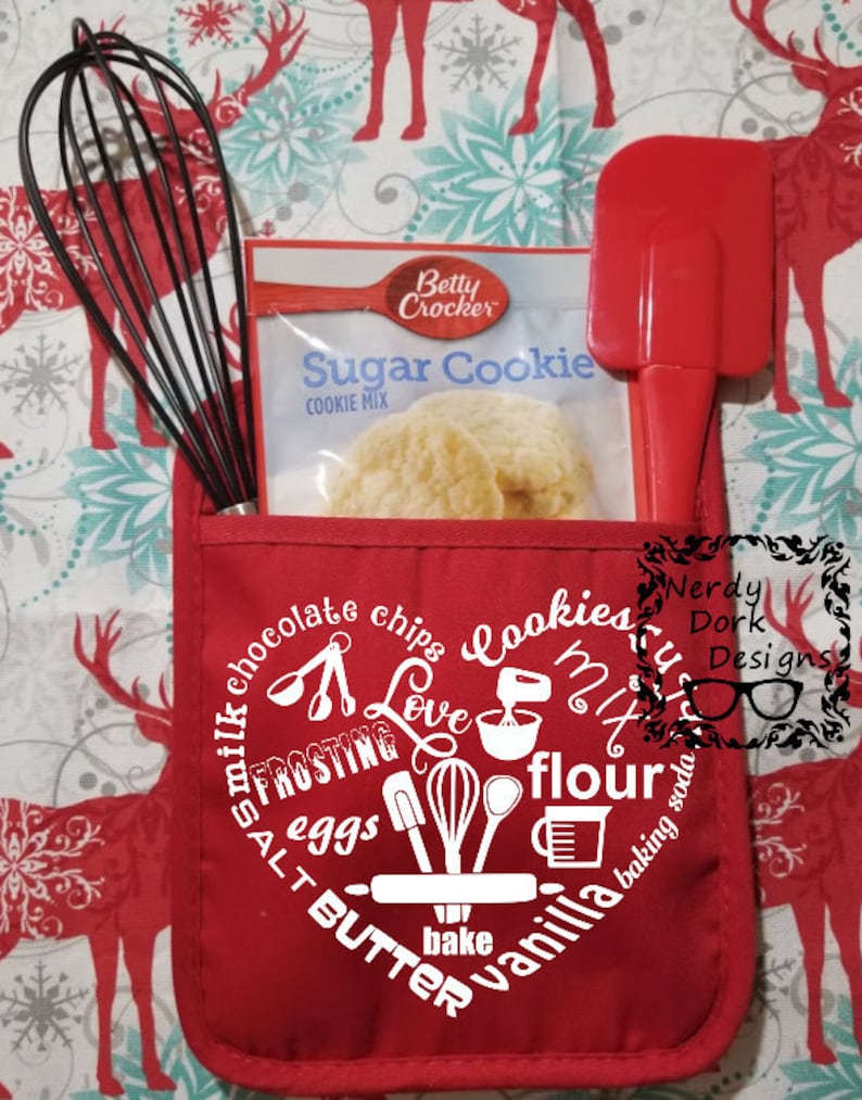 Op de afbeelding: Een rode ovenwant met een wit hartontwerp met woorden en pictogrammen die te maken hebben met bakken, waaronder "cookies", "love", "flour", "butter", "vanilla" en "bake". Op de achtergrond zijn een zak Betty Crocker Sugar Cookie mix, een rode spatel en een garde te zien.