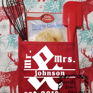 Op de afbeelding: Een rode ovenwant met een wit ontwerp dat "mr. & mrs. Johnson est. 2018" zegt met een decoratieve versiering. De ovenwant heeft een zakje met een pakje suikerkoekjesmix en een rode spatel.