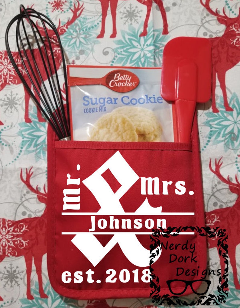 Op de afbeelding: Een rode ovenwant met een wit ontwerp dat "mr. & mrs. Johnson est. 2018" zegt met een decoratieve versiering. De ovenwant heeft een zakje met een pakje suikerkoekjesmix en een rode spatel.