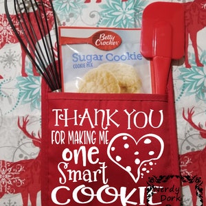 Op de afbeelding: Een rode ovenwant met witte tekst die zegt "Bedankt dat je me een slimme bakker hebt gemaakt" met een hartvorm met witte stippen. De ovenwant heeft een zakje met een garde, spatel en een zak Betty Crocker Sugar Cookie mix.