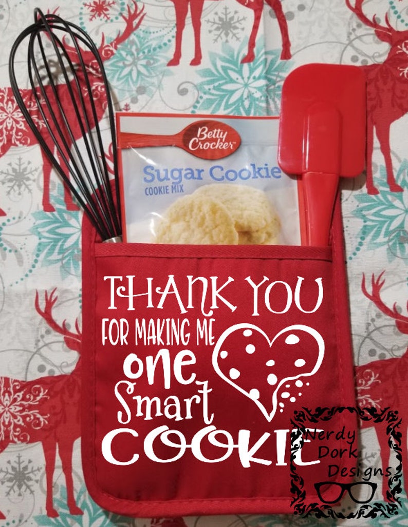 Op de afbeelding: Een rode ovenwant met witte tekst die zegt "Bedankt dat je me een slimme bakker hebt gemaakt" met een hartvorm met witte stippen. De ovenwant heeft een zakje met een garde, spatel en een zak Betty Crocker Sugar Cookie mix.