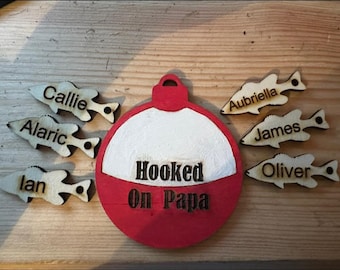 Op de afbeelding: Een rood en wit houten ornament in de vorm van een dobber met de tekst "Hooked on Papa" op het rode gedeelte. Vijf houten visornamenten met namen erop omringen de dobber: Callie, Alaric, Ian, Aubriella, James en Oliver.