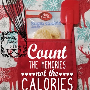 Op de afbeelding: Een rode ovenwant met de tekst "Count the memories not the calories" in wit. De ovenwant heeft een zakje met een garde, een spatel en een pakje suikerkoekmix.