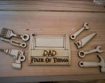 Op de afbeelding: Houten gereedschapskist met de woorden "Dad Fixer of Things" en verschillende gereedschappen, waaronder een zaag, een sleutel, een hamer, een kwast en een boor. Elk gereedschap heeft een naam: Ian, James, Oliver.