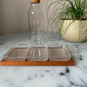 K&ouml;nnte beinhalten: Ein Satz von vier klaren Glasschalen mit Holzgriffen, angeordnet auf einem Holztablett. Die Schalen befinden sich auf einer wei&szlig;en Marmorfl&auml;che. Eine klare Glasflasche mit einem Korkverschluss befindet sich im Hintergrund.