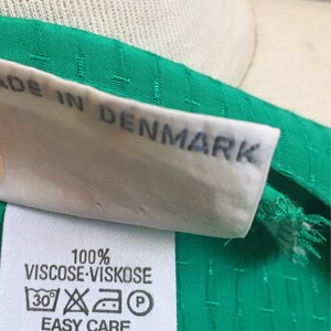 Puede incluir: Una etiqueta de tela blanca con el texto "100% Viscose Viskose" e instrucciones de cuidado. La etiqueta est&aacute; unida a una tela verde con un patr&oacute;n texturizado. La etiqueta tambi&eacute;n dice "Easy Care" y "Made in Denmark".