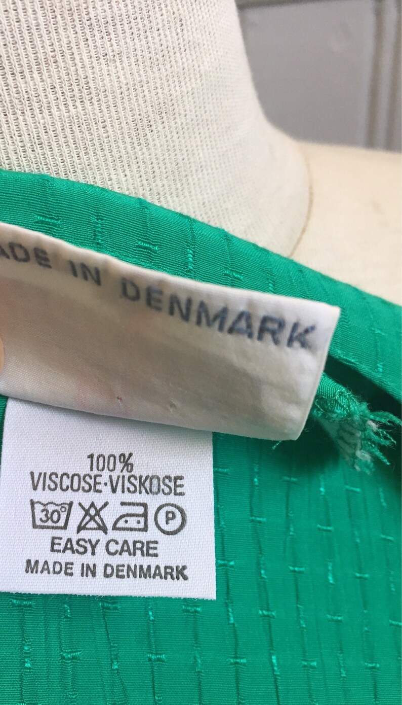 Puede incluir: Una etiqueta de tela blanca con el texto "100% Viscose Viskose" e instrucciones de cuidado. La etiqueta est&aacute; unida a una tela verde con un patr&oacute;n texturizado. La etiqueta tambi&eacute;n dice "Easy Care" y "Made in Denmark".
