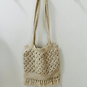 May include: A beige crocheted tote bag with fringe hanging from the bottom. The bag has a long strap and is hanging on a wooden rack.