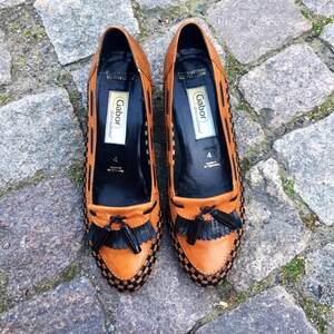 May include: A pair of brown leather loafers with black tassels. The loafers have a black and brown woven pattern and the brand name "Gabor International" is printed on the inside of each shoe.