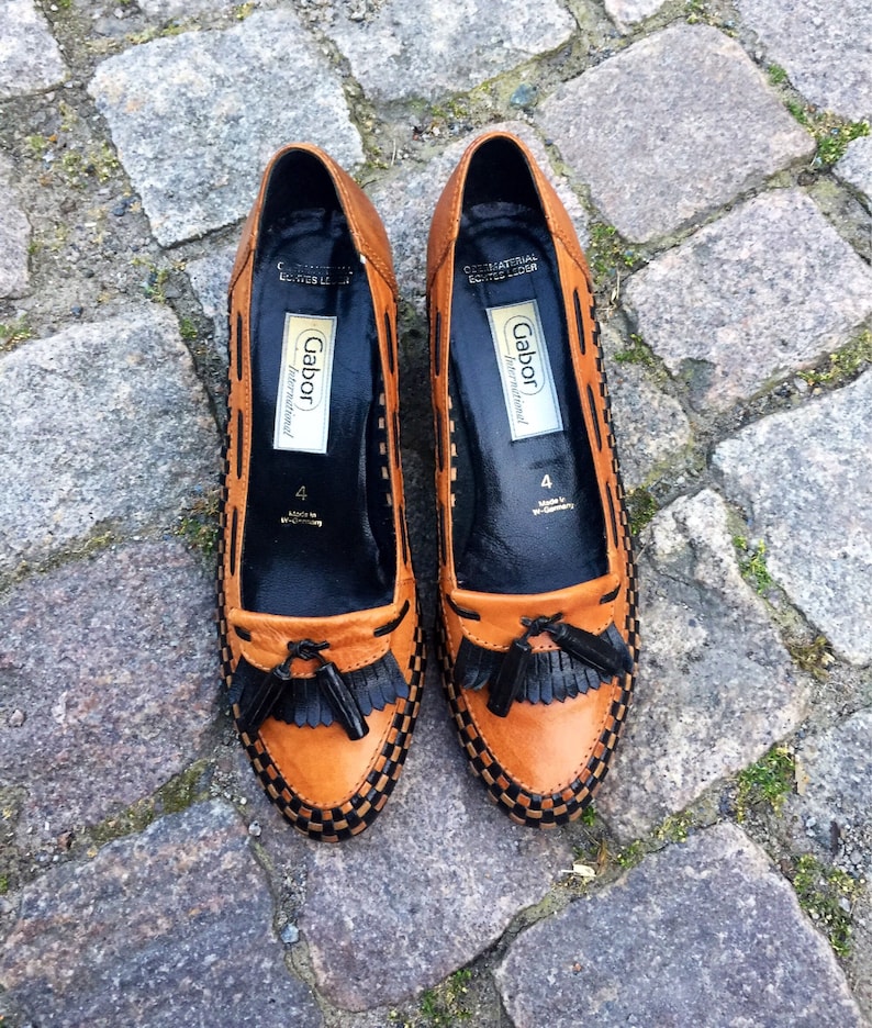 May include: A pair of brown leather loafers with black tassels. The loafers have a black and brown woven pattern and the brand name "Gabor International" is printed on the inside of each shoe.