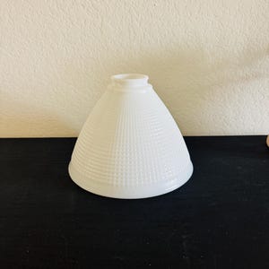 May include: A white, cone-shaped lampshade with a textured, grid-like pattern. The lampshade has a wide base that tapers to a smaller opening at the top. The lampshade is made of opaque white glass or ceramic.