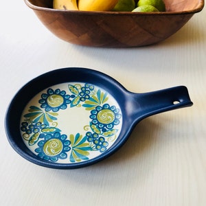 May include: A blue ceramic dish with a white center and a floral design in shades of green and blue. The dish has a handle on the side.