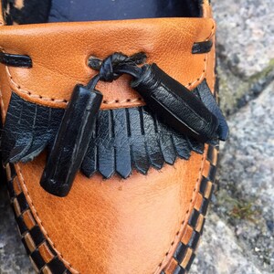 May include: Close-up of a brown leather loafer with black leather trim and tassels. The loafer has a woven pattern around the edge and says "Made in W-Germany" and "4" on the inside.