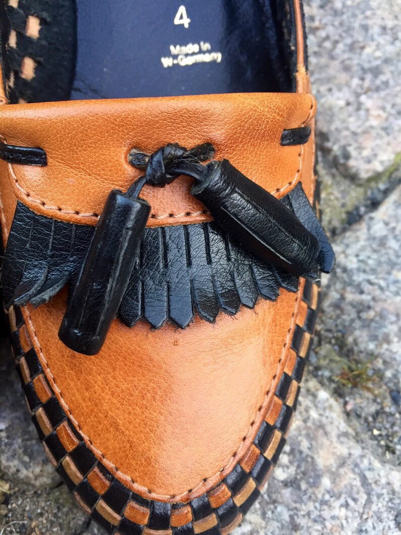 May include: Close-up of a brown leather loafer with black leather trim and tassels. The loafer has a woven pattern around the edge and says "Made in W-Germany" and "4" on the inside.