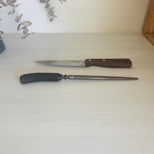 May include: Two kitchen knives on a white surface. The top knife has a stainless steel blade and a dark brown wooden handle. The bottom knife is a steel honing steel with a dark handle. The knives are for food preparation.