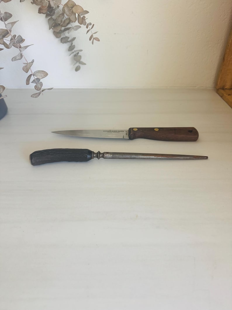 May include: Two kitchen knives on a white surface. The top knife has a stainless steel blade and a dark brown wooden handle. The bottom knife is a steel honing steel with a dark handle. The knives are for food preparation.