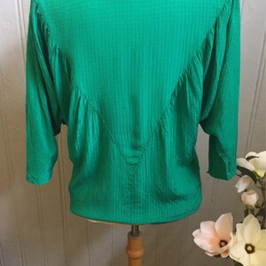 Puede incluir: Una blusa verde de manga larga con textura, cuello en V y un detalle fruncido en la espalda.