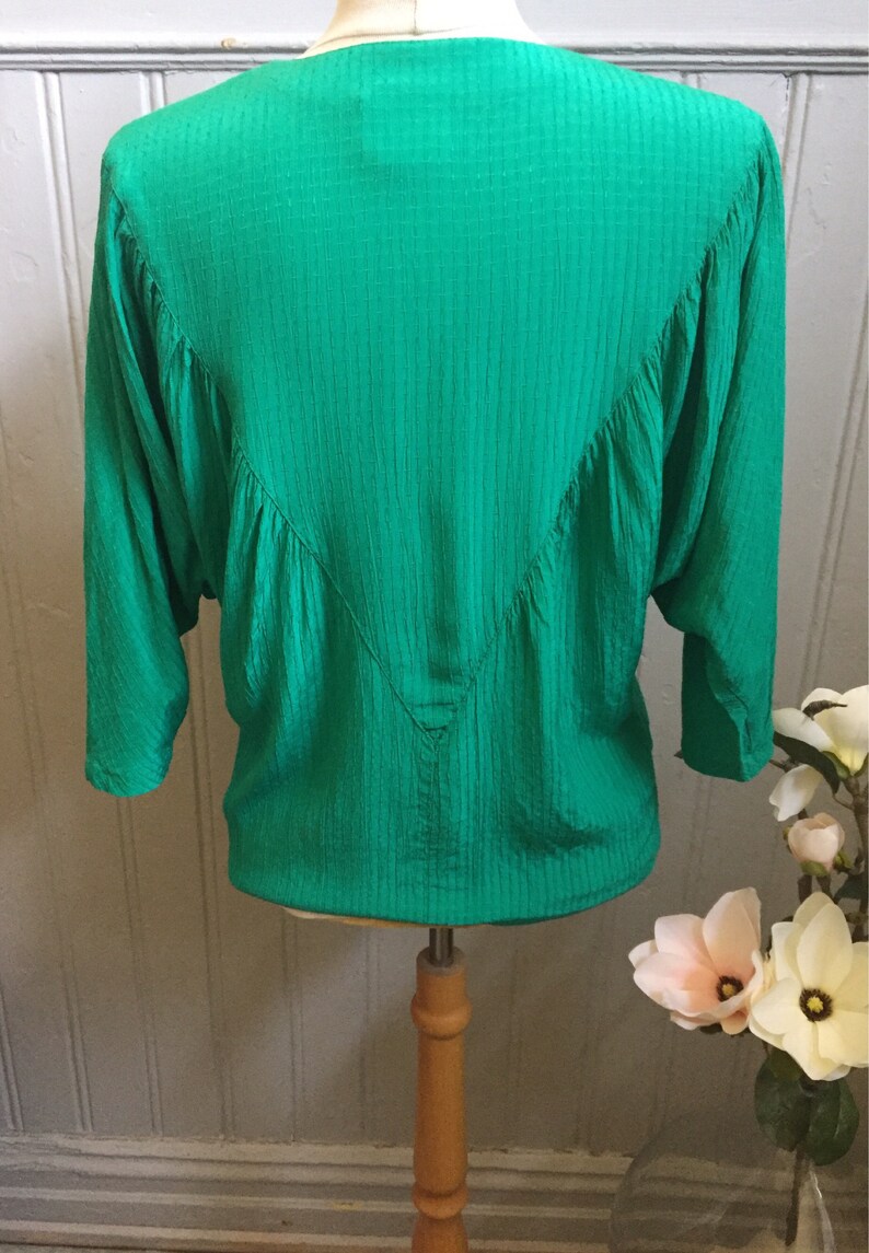 Puede incluir: Una blusa verde de manga larga con textura, cuello en V y un detalle fruncido en la espalda.