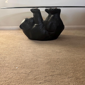 May include: A black bear-shaped coffee table with a clear glass top. The bear is lying on its back with its paws supporting the glass. The table is a unique piece of furniture.