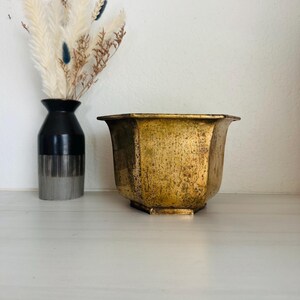 May include: A hexagonal brass planter with a weathered, mottled gold finish and a wide, flared rim. The metal surface displays a textured, aged patina with darker spots throughout, and the base is slightly tapered with a flat, hexagonal bottom.