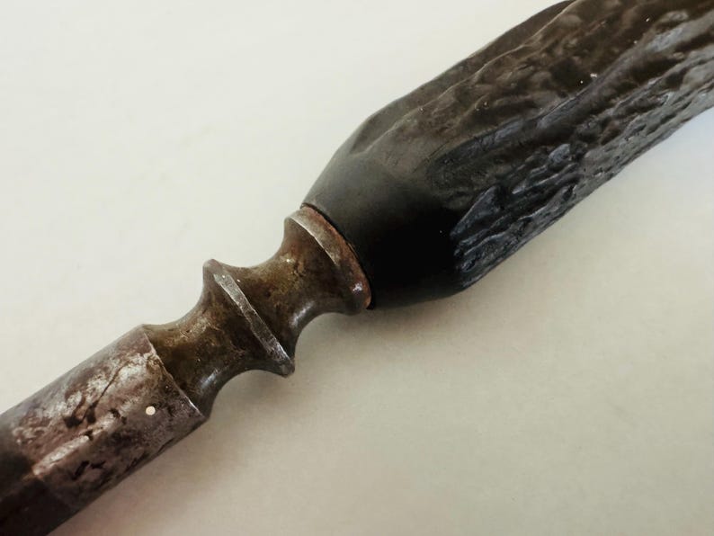 May include: Close-up of a vintage knife. The handle is dark brown and textured, leading to a black section near the blade. The blade's base is a rusty, aged metal. The knife has a decorative, ornate design.