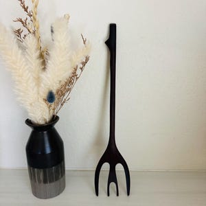 May include: A dark brown wooden serving fork with a unique, artistic design. The fork has three tines and a long, slender handle. The handle has a decorative element at the top. The fork is displayed next to a black vase with dried flowers.