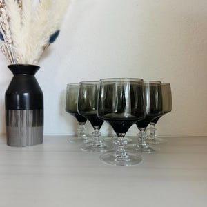 May include: Six gray-tinted wine glasses with clear stems and bases are arranged on a white surface. A black and silver vase with dried flowers is to the left. The glasses have a classic wine glass shape.