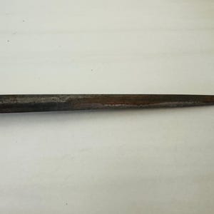 May include: A close-up of a metal tool with a tapered, pointed end. The tool has a dark, weathered appearance with areas of rust and discoloration, suggesting age and use. The background is a plain, light surface.