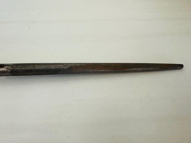 May include: A close-up of a metal tool with a tapered, pointed end. The tool has a dark, weathered appearance with areas of rust and discoloration, suggesting age and use. The background is a plain, light surface.