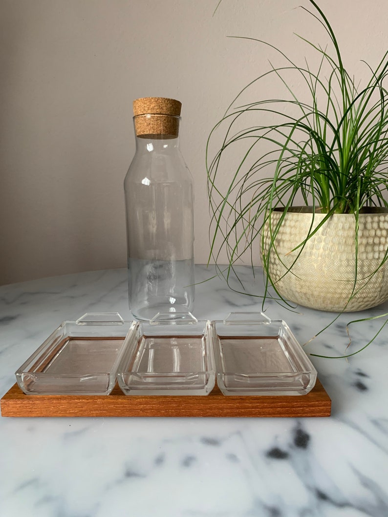K&ouml;nnte beinhalten: Ein Satz von vier klaren Glasschalen mit Holzgriffen, angeordnet auf einem Holztablett. Die Schalen befinden sich auf einer wei&szlig;en Marmorfl&auml;che. Eine klare Glasflasche mit einem Korkverschluss befindet sich im Hintergrund.