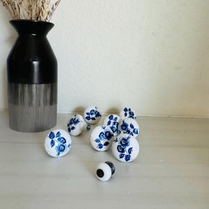 May include: A collection of white ceramic cabinet knobs with a blue floral design. The knobs are scattered on a light-colored surface, with a black and silver vase holding dried flowers in the background.