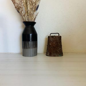 May include: A black and silver vase with dried flowers and a rusty metal bell with a handle. The vase has a black top and a silver striped bottom. The bell is rectangular and has a handle on top.